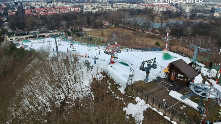 Narciarze zjeżdżają po sztucznie naśnieżonym stoku, otoczonym przez budynki miejskie i drzewa.