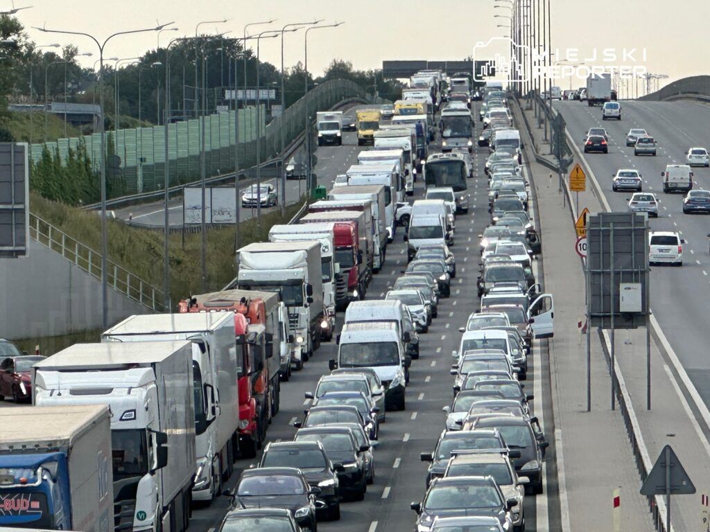 Gęsty korek samochodowy na dwupasmowej drodze, z dużą ilością ciężarówek i osobówek, w tle widoczne znaki drogowe.
