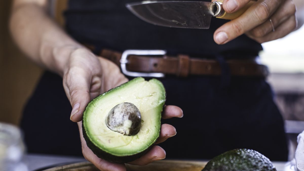 Salad Preparation: Slicing Avocado 1A woman prepares to slice an avocado using a safe technique, protecting her fingers and using a stainless steel knife on a wooden chopping board.