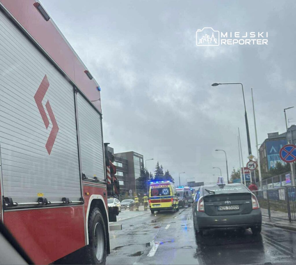 Czerwony wóz strażacki stoi na drodze, obok niego karetka z włączonymi sygnałami świetlnymi, w deszczowej aurze.