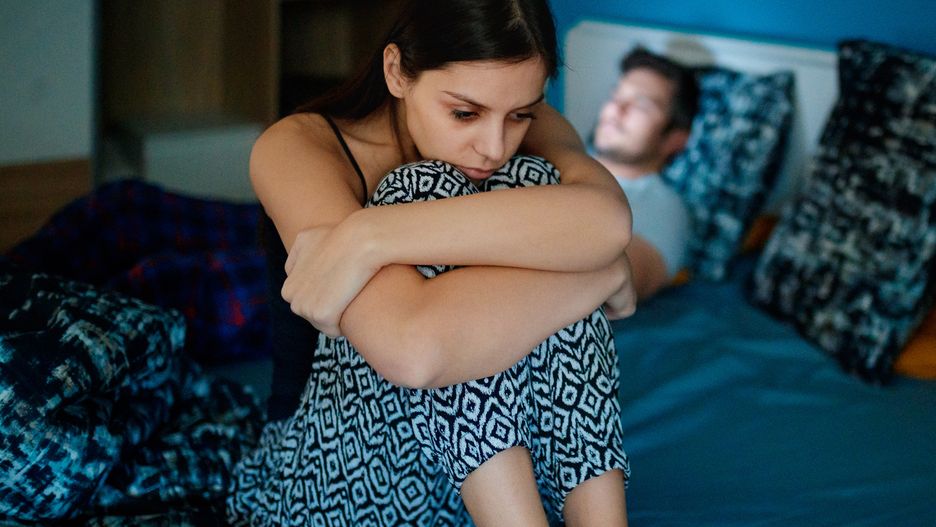 Sad woman sitting on bed with partner in backgroundSad girlfriend sitting on bed edge, holding her legs after fight, her partner is lying in bed in background, his face is lit by smart phonezoranm