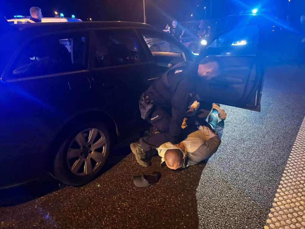 Policjant obezwładnia mężczyznę leżącego na drodze obok zaparkowanego samochodu w nocy, w otoczeniu radiowozów.