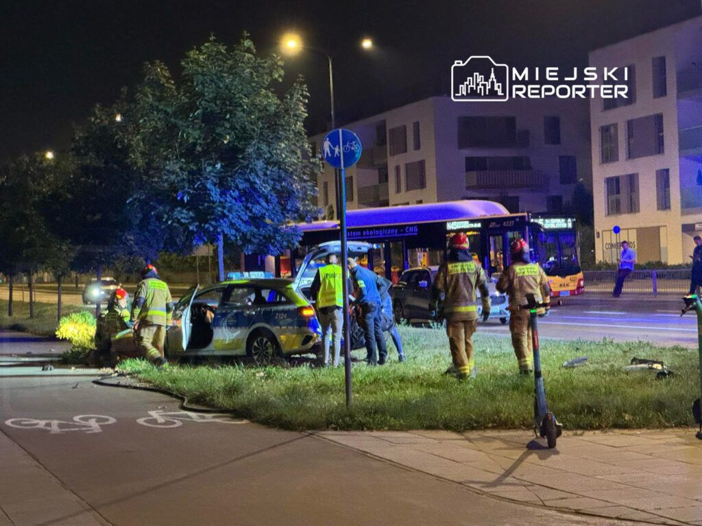 Grupa strażaków udziela pomocy przy wypadku drogowym, w tle widoczny autobus i oświetlone budynki.