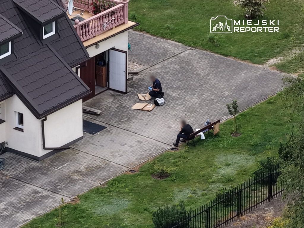 Dwóch mężczyzn siedzących na ławce, jeden z nich przegląda dokumenty na ziemi, w tle dom jednorodzinny.