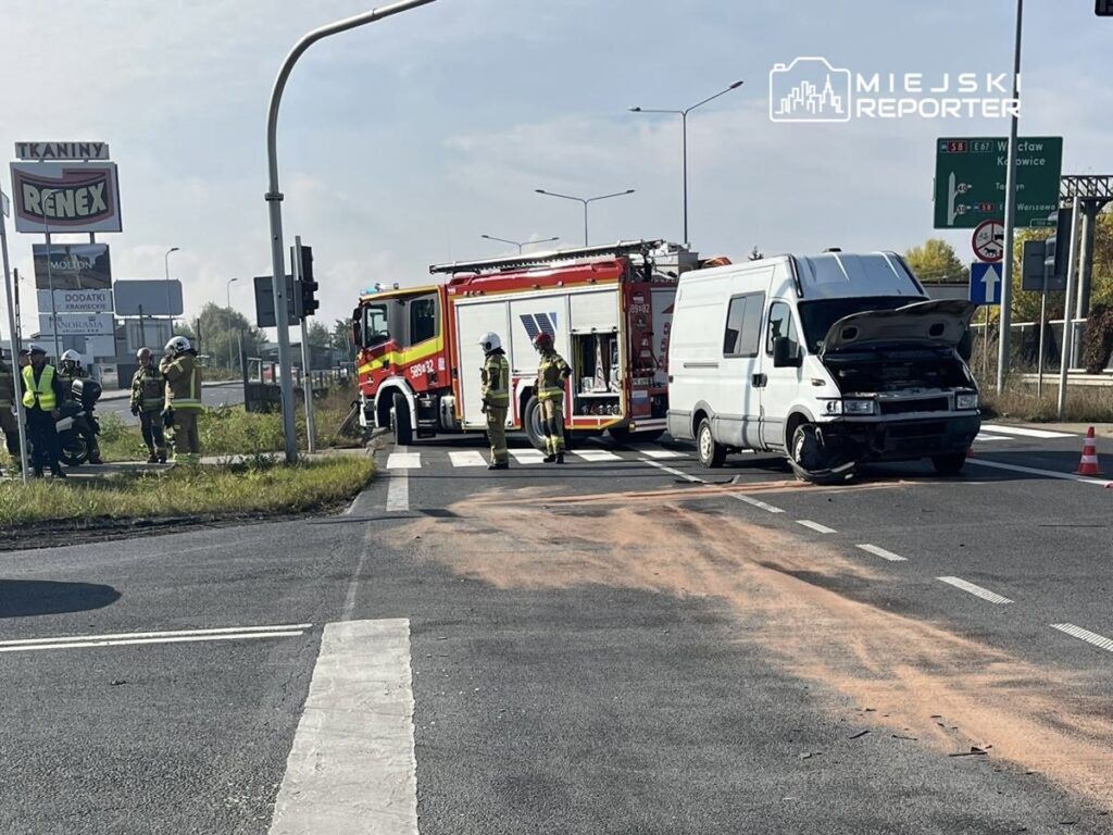 Na skrzyżowaniu stoi uszkodzony biały pojazd dostawczy z otwartą maską, obok pracują strażacy w mundurach.