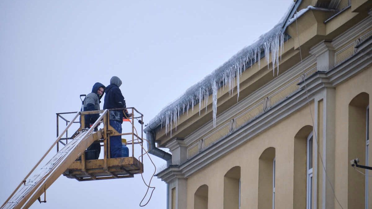 mężczyźni w podnośniku przy pomocy palnika usuwają zwisające sople lodu
