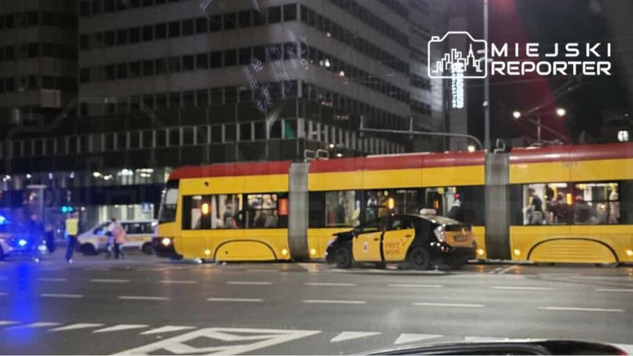 Tramwaj z czerwono-żółtym nadwoziem stoi na skrzyżowaniu, obok policyjne radiowozy i taksówka zaparkowana na jezdni.