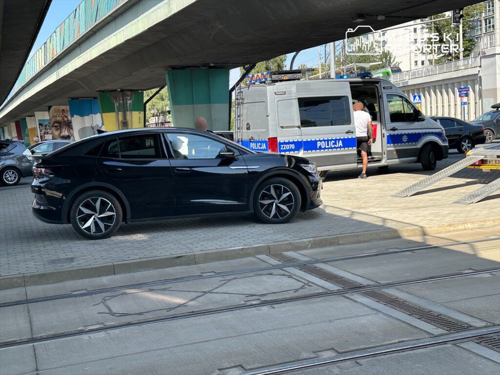 Czarny samochód osobowy zaparkowany obok radiowozu policyjnego, w tle widoczne graffiti na murze.