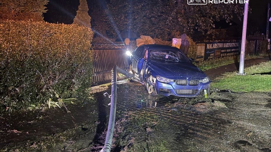 Niebieski samochód osobowy stoi na poboczu drogi, obok widoczna jest woda i uszkodzona brama.