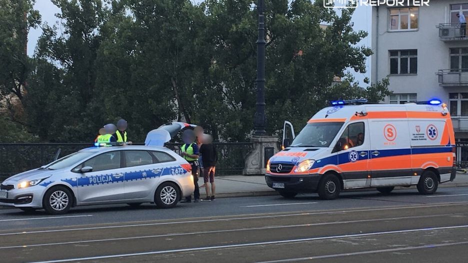 Policjanci w kamizelkach odblaskowych rozmawiają z mężczyzną na moście obok radiowozu i karetki pogotowia.