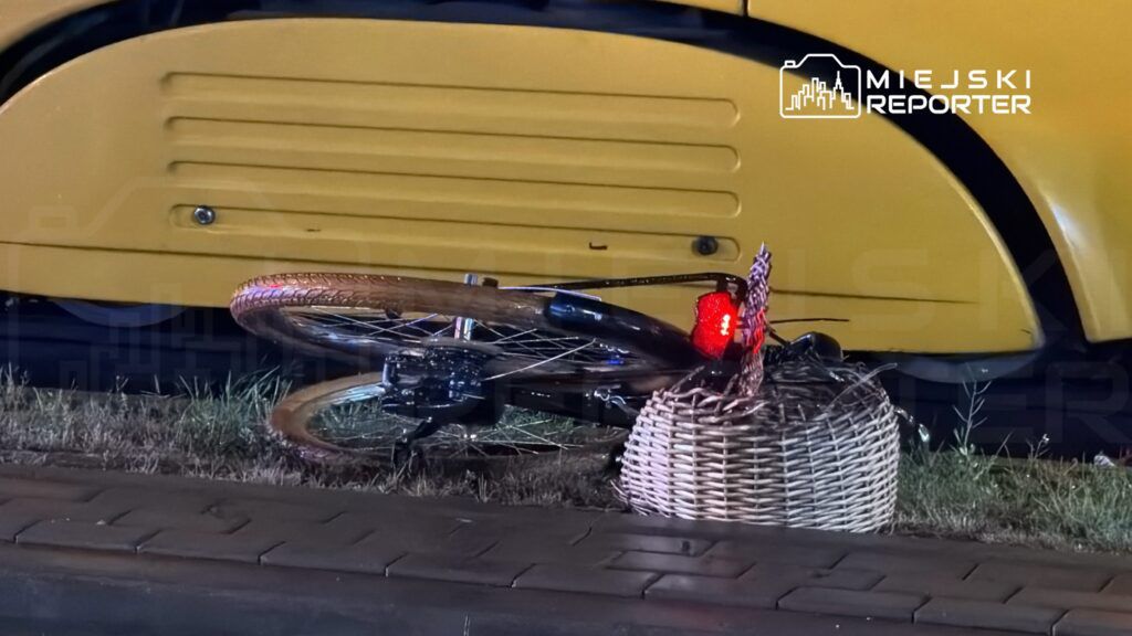 Złamany rower leży na trawie obok żółtego pojazdu, widoczna czerwona lampka na tylnym kole.