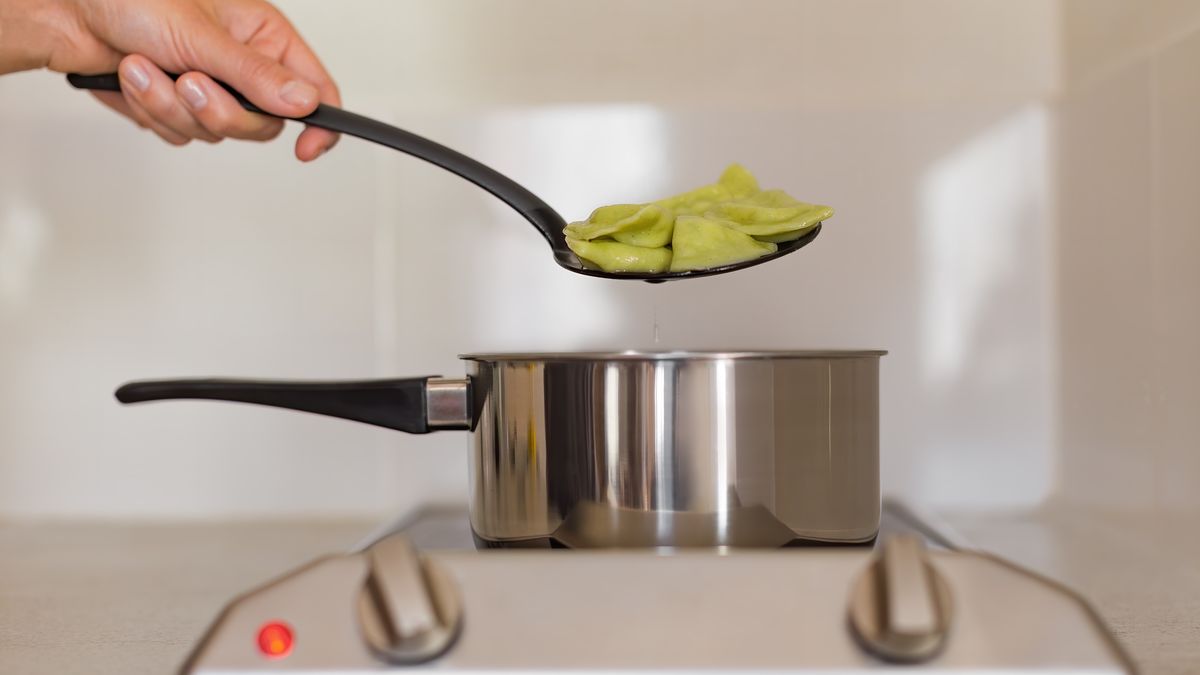 A hand with a slotted spoon takes dumplings out of the pot.A hand with a hollowed spoon takes out green vegetarian dumplings from the ladle, which is standing on the stovePavel Adashkevich