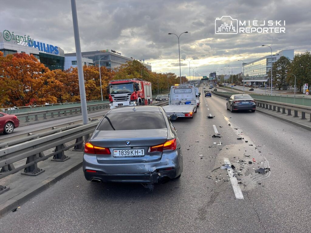 Na jezdni znajduje się uszkodzony samochód osobowy oraz resztki zderzenia, obok przejeżdża wóz strażacki. W tle widać nowoczesne budynki i kolorowe drzewa.