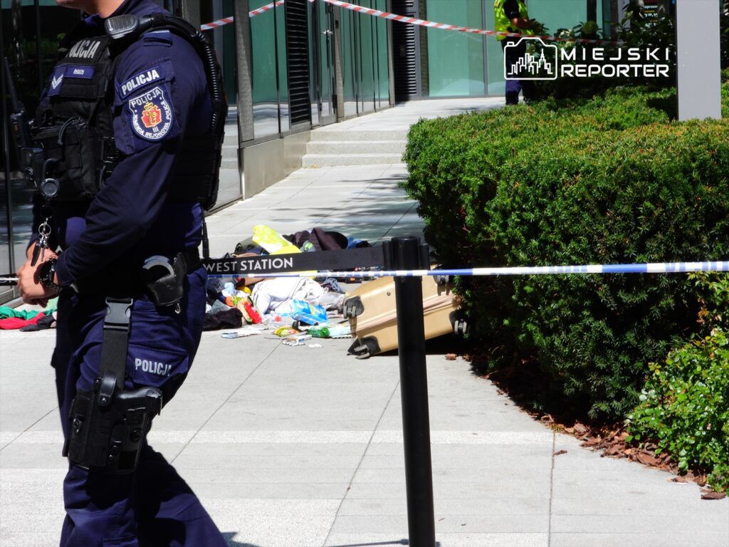 Policjant w mundurze stoi przy taśmie zabezpieczającej, obok leżą różne przedmioty i walizka.
