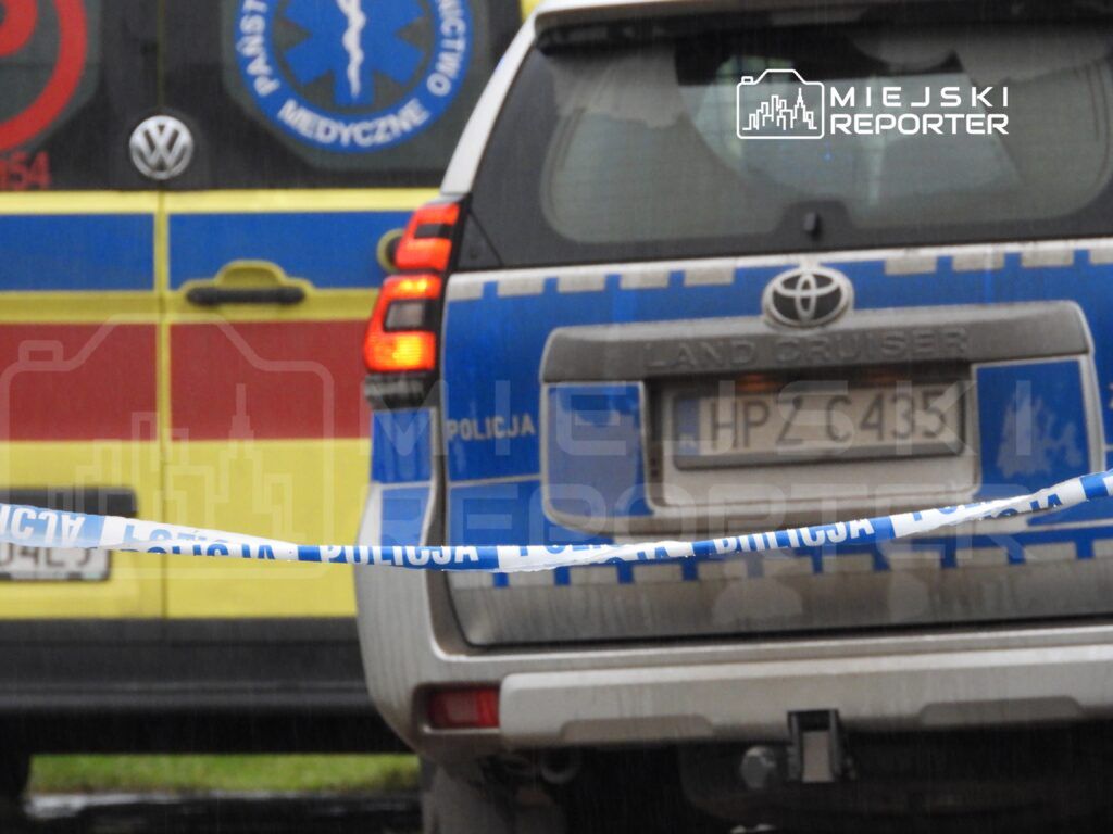 Policyjny radiowóz zaparkowany za taśmą zabezpieczającą, w tle karetka pogotowia.