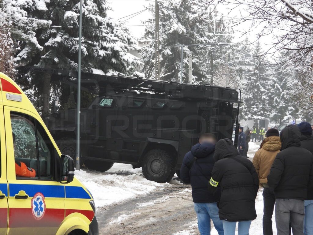 Grupa osób w zimowych kurtkach obserwuje policyjny pojazd opancerzony na zaśnieżonej ulicy. W tle widoczne drzewa pokryte śniegiem.