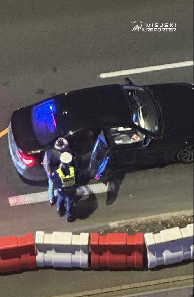 Policyjny funkcjonariusz w kamizelce odblaskowej rozmawia z kierowcą stojącego czarnego samochodu.