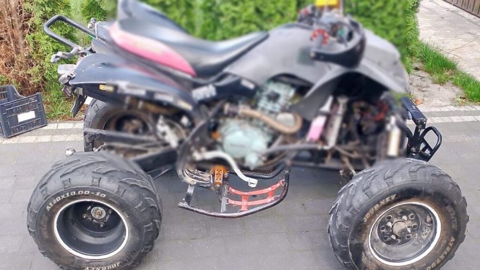 Czarny quad z dużymi oponami stoi na kostce brukowej, w tle widoczne krzewy i ogrodzenie.
