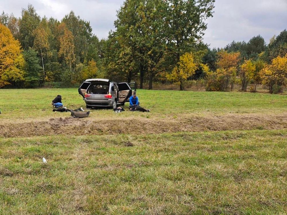 Dwóch mężczyzn w dresach pracuje przy samochodzie na otwartej przestrzeni, w tle drzewa z kolorowymi liśćmi.