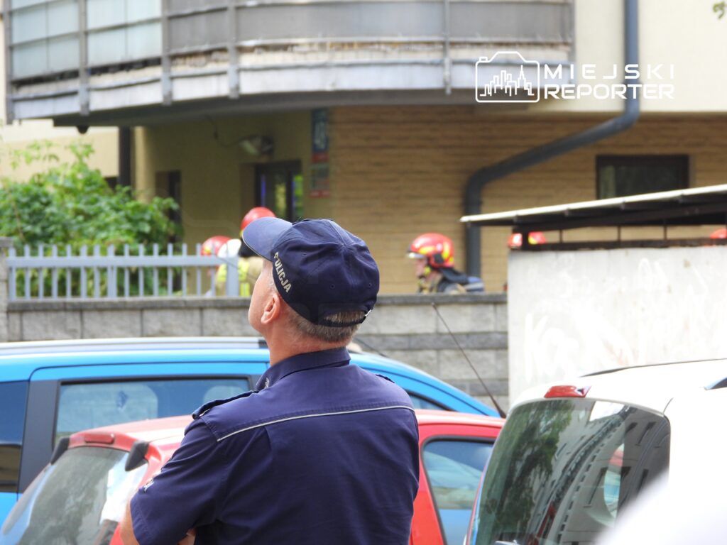 Mężczyzna w mundurze policyjnym obserwujący sytuację przed budynkiem, w tle strażacy w hełmach.