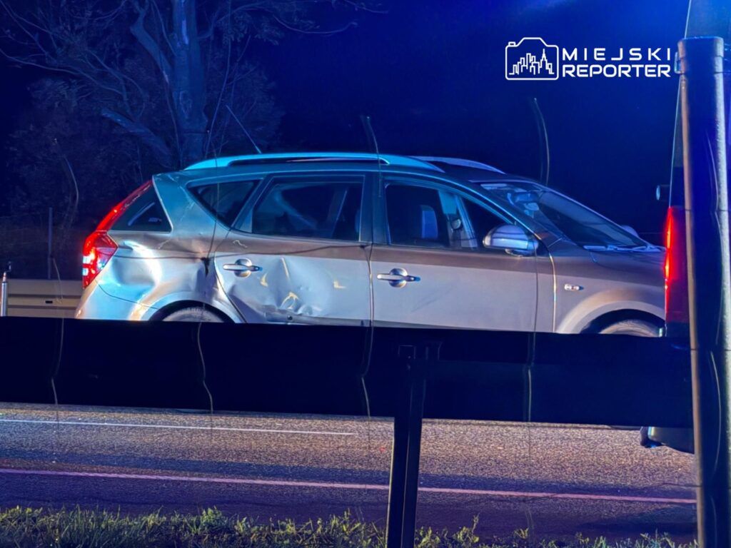 Uszkodzony srebrny samochód osobowy zatrzymany na poboczu drogi, w tle niebieskie światła policyjne.