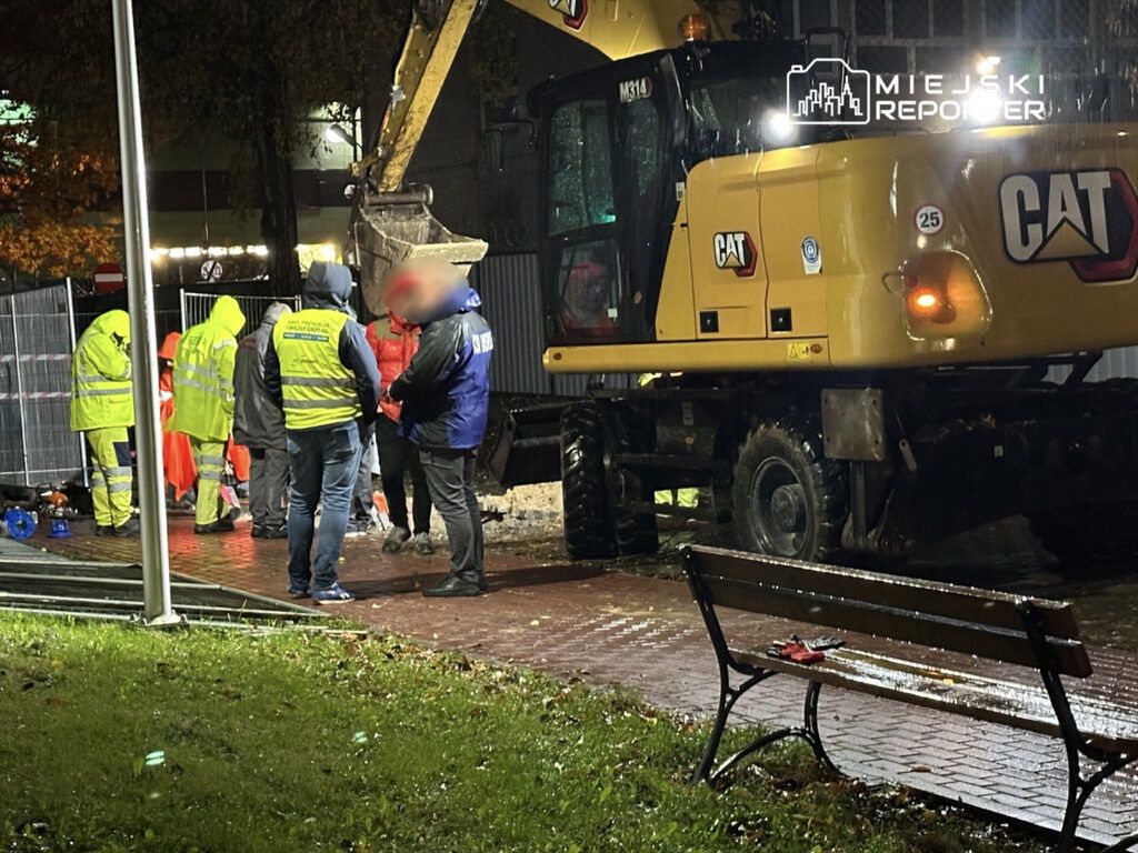Grupa pracowników w odblaskowych kurtkach rozmawia w deszczowej scenerii obok żółtej koparki.