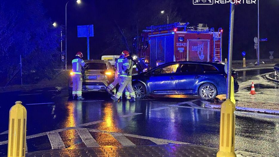 Trzech strażaków w kaskach bada uszkodzone samochody na mokrej jezdni w nocy, obok wozu strażackiego.