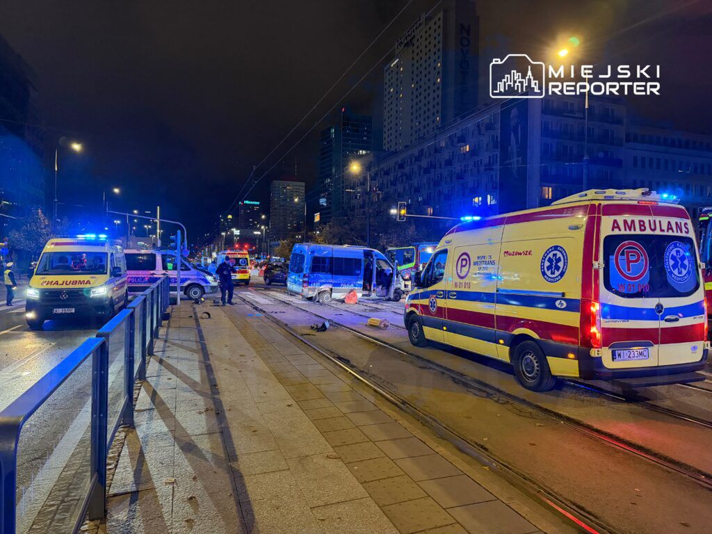 Na ulicy, w nocy, zespoły ratunkowe i policja interweniują po wypadku drogowym. W tle widać nowoczesne budynki.