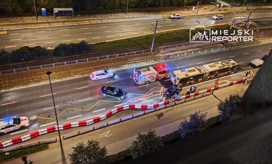 W nocy widać wypadek drogowy z udziałem samochodu osobowego i autobusu, otoczonego przez pojazdy służb ratunkowych.