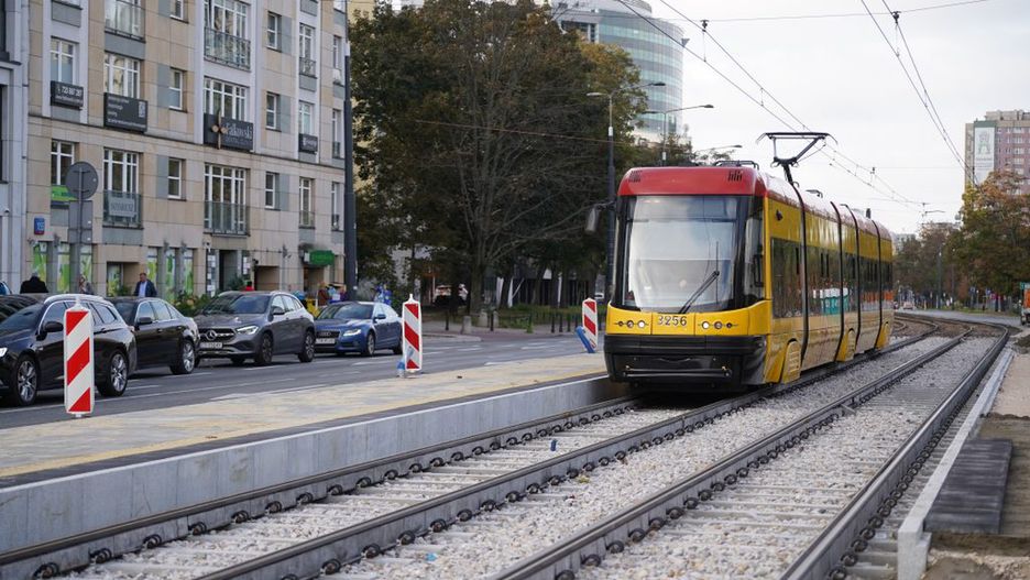 Tramwaj w żółto-czerwonym kolorze zbliża się do przystanku, obok ruchliwa ulica z zaparkowanymi samochodami.