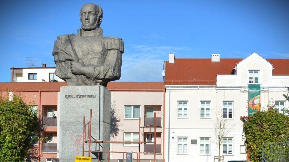 Pomnik gen. Józefa Bema w otoczeniu budynków mieszkalnych, z widocznymi zabezpieczeniami budowy i armatą w tle.