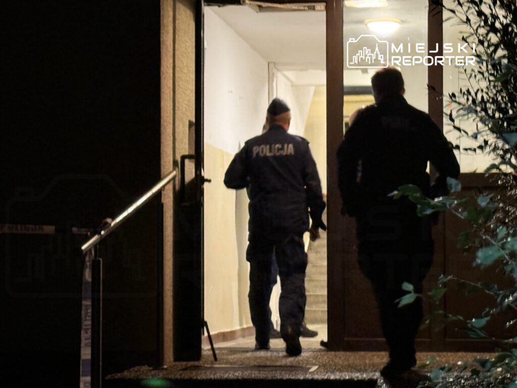 Dwóch policjantów w mundurach wchodzi do budynku przez otwarte drzwi w nocy.