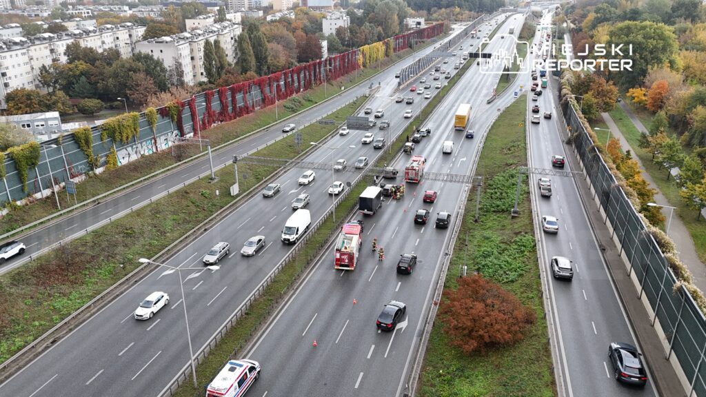 Na jezdni autostrady zatrzymane pojazdy, w tym ciężarówka, a obok pracują strażacy i ratownicy. W tle widoczne bloki mieszkalne.