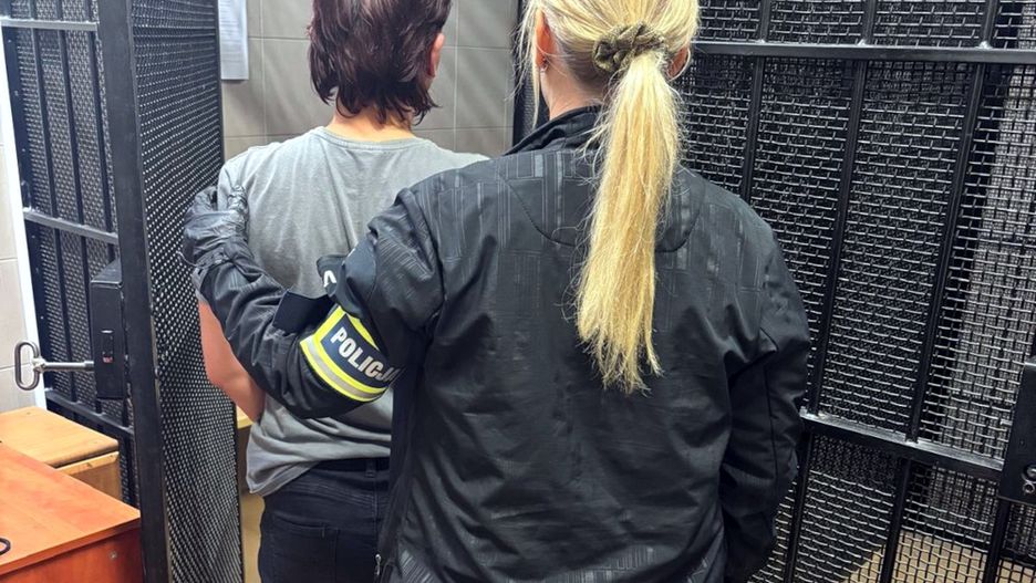 Kobieta w mundurze policyjnym prowadzi osobę w szarej koszulce przez metalowe drzwi w budynku policyjnym.