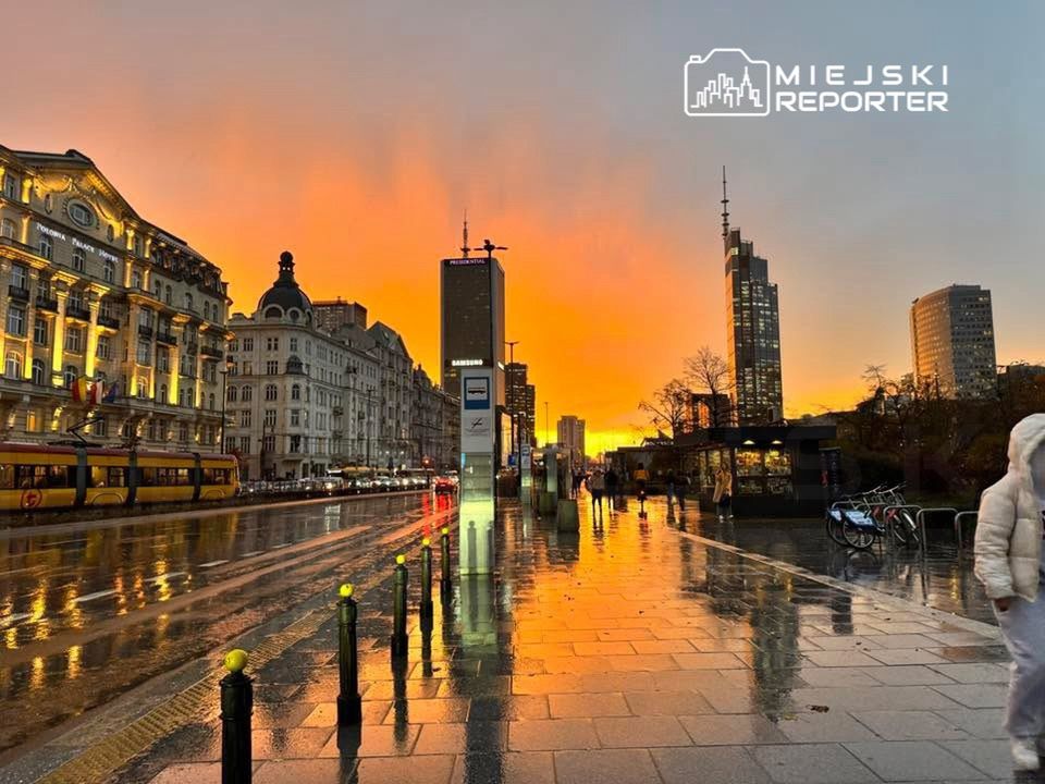 Uliczka w mieście odbijająca kolorowy zachód słońca na mokrej nawierzchni, z tramwajem i nowoczesnymi budynkami w tle.