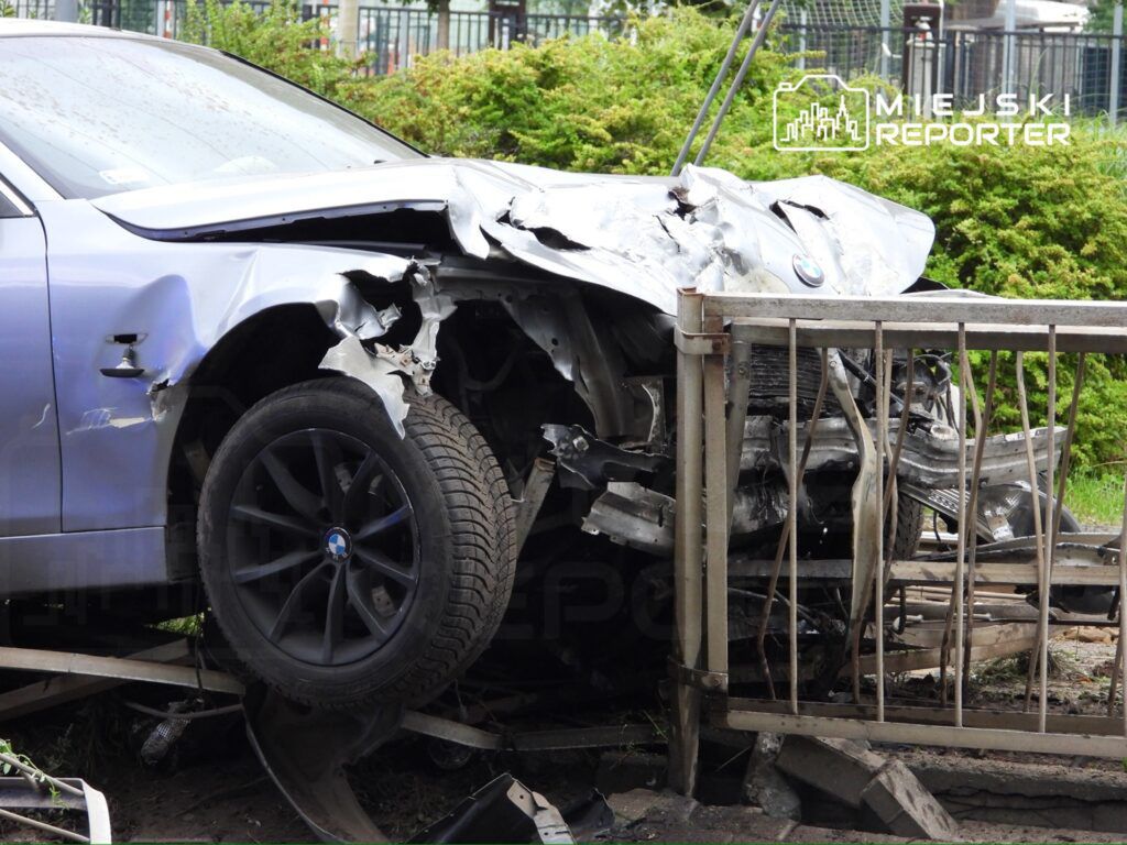 Uszkodzony samochód osobowy marki BMW wjeżdżający w ogrodzenie, z widocznymi zniszczeniami przodu i koła.