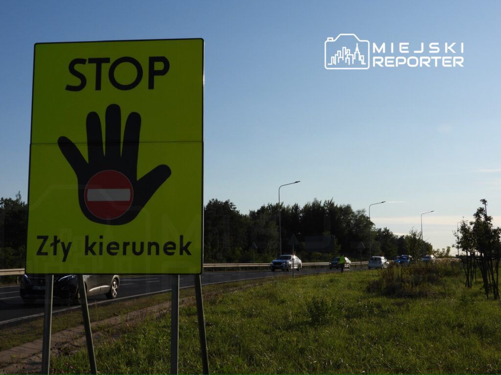 Duży, żółty znak drogowy z napisem "STOP" i symbolem ręki, informujący o złym kierunku, przy ruchliwej drodze.