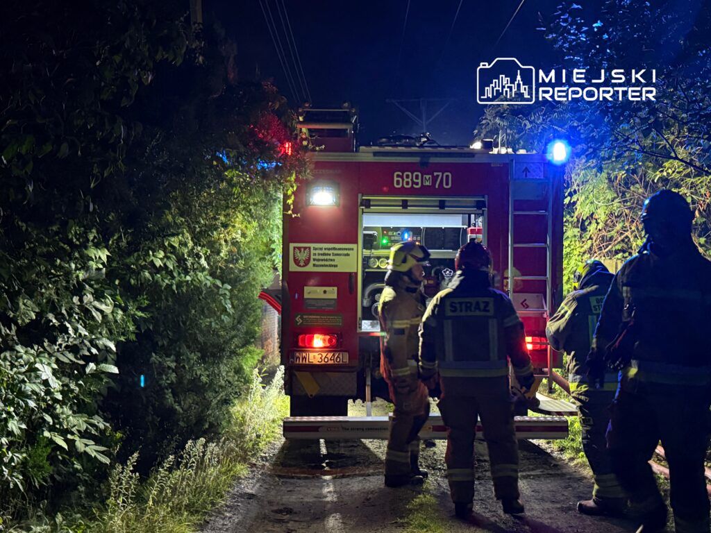 Strażacy w mundurach omawiają sytuację obok wozu strażackiego na wąskiej, oświetlonej drodze w nocy.
