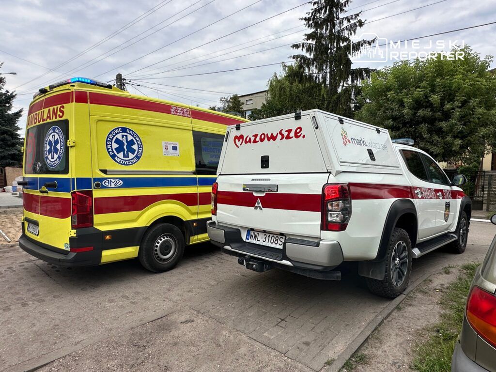 Ambulans i pojazd ratunkowy stoją na ulicy, obok drzew i budynków mieszkalnych. Na ambulansie widoczne logo Państwowego Ratownictwa Medycznego.