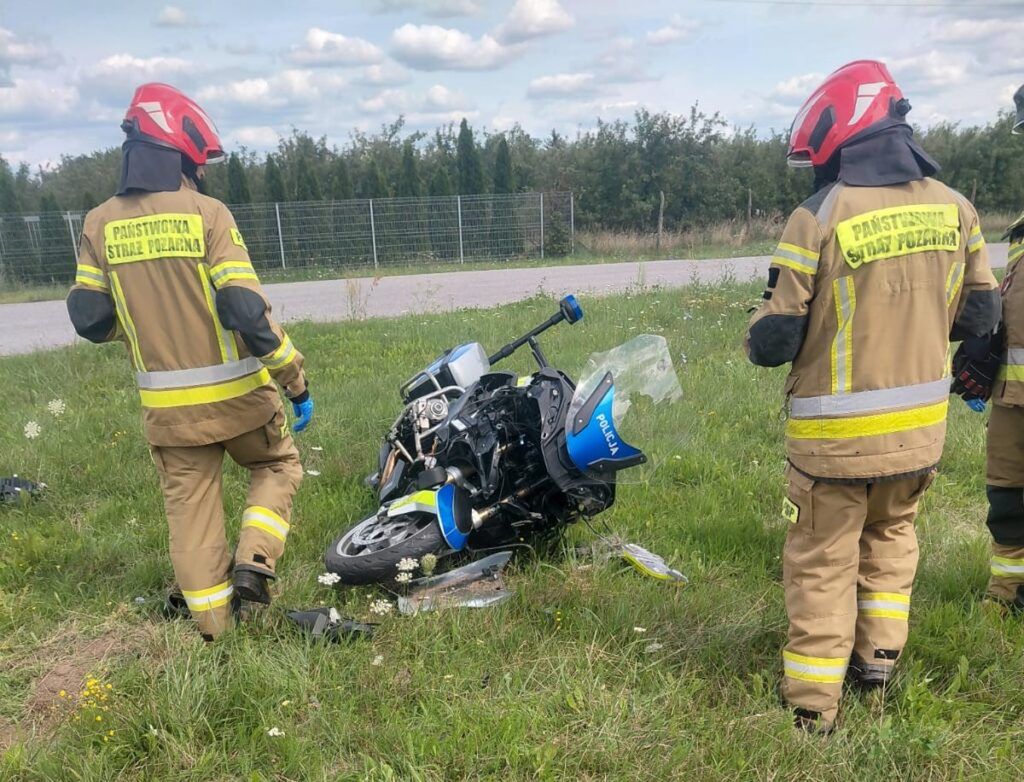 Strażacy w hełmach oceniają przewrócony motocykl na poboczu drogi, w tle zieleń i ogrodzenie.