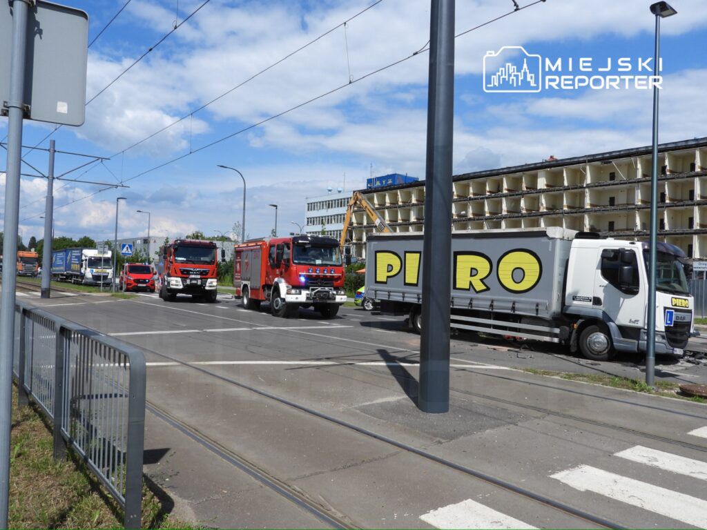 Czerwone wozy strażackie oraz ciężarówki stojące na skrzyżowaniu, obok modernizowanego budynku.