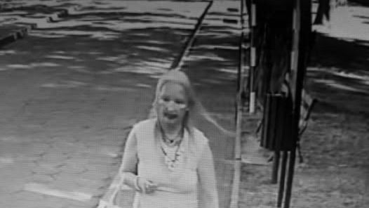 Kobieta w jasnym stroju spacerująca po chodniku w parku, trzymająca torebkę.