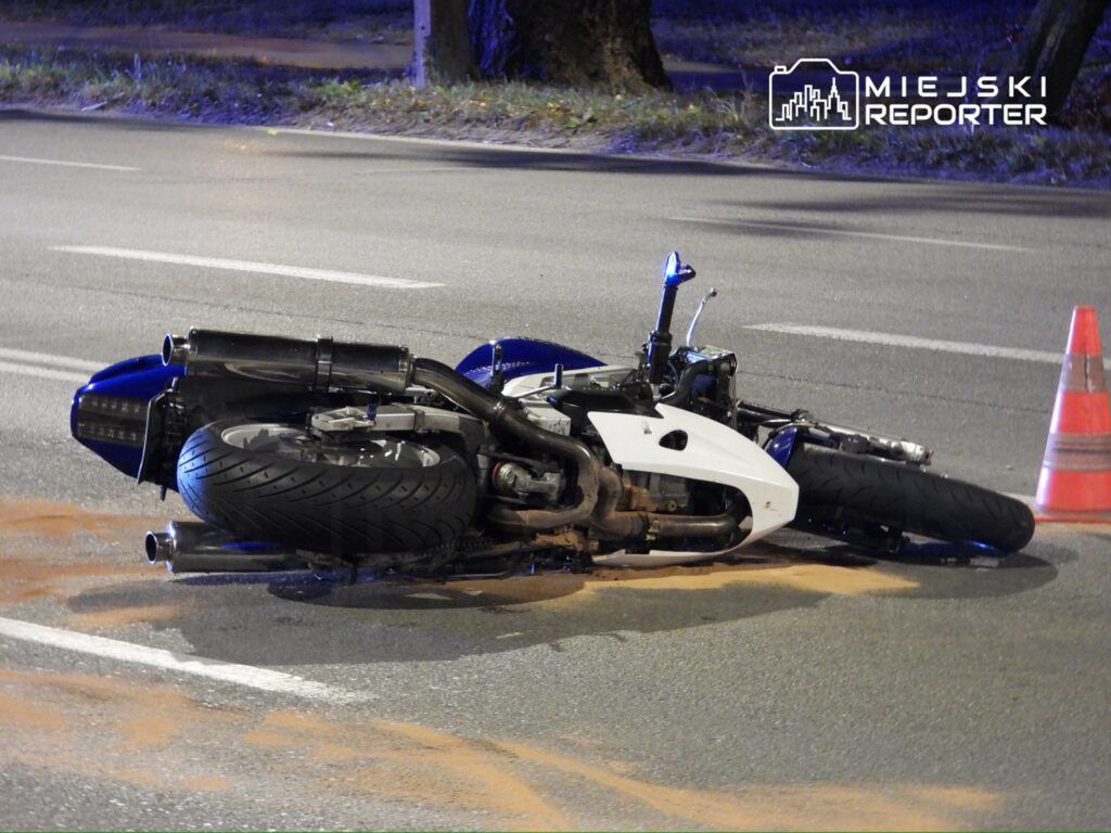 Motocykl leży na jezdni, obok pomarańczowy trójkąt ostrzegawczy, a na asfalcie widoczna plama.