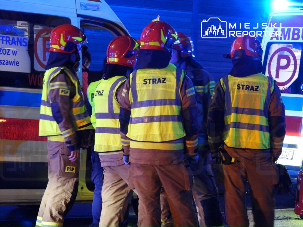 Grupa strażaków w hełmach i odblaskowych kamizelkach omawia sytuację w pobliżu karetki pogotowia.