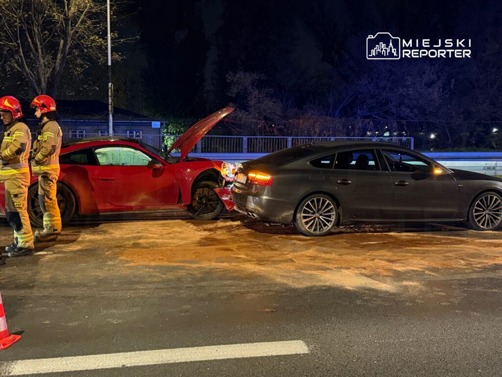 Dwóch strażaków w uniformach stoi obok czerwonego sportowego auta z otwartą maską, obok czarnego samochodu po kolizji na drodze.