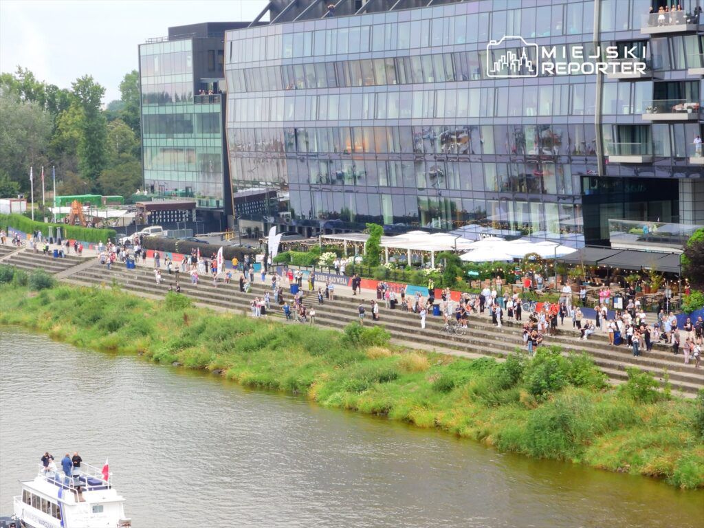 Grupa ludzi spacerująca wzdłuż brzegu rzeki obok nowoczesnego biurowca z przeszkloną fasadą.