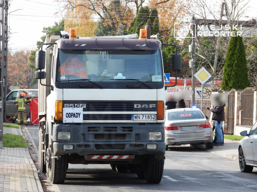 W tle widoczny ciężarówka z napisem "ODPADY", a obok strażacy pracujący przy samochodzie osobowym. Policja monitoruje sytuację.