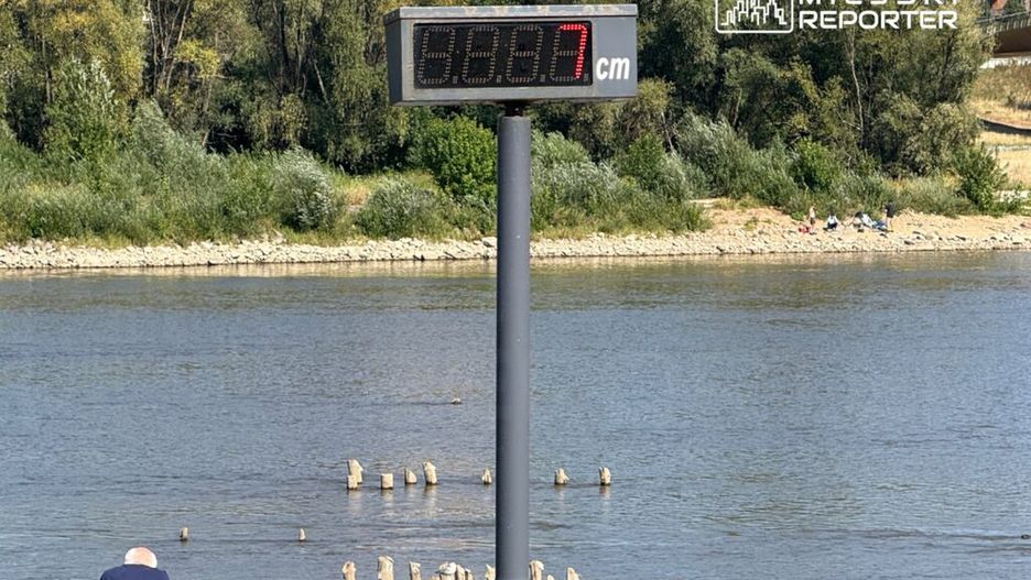 Dziecko w różowej koszulce i starszy mężczyzna obserwują rzekę, obok wyświetlacz z wysokością wody 68,7 cm.