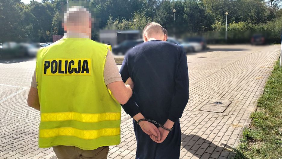 Mężczyzna w kajdankach prowadzony przez policjanta w kamizelce na parkingu otoczonym drzewami.