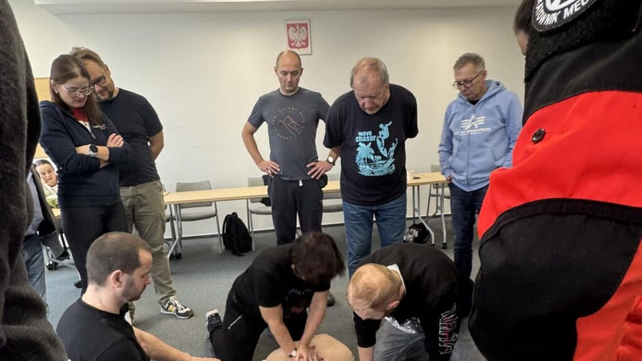 Grupa osób obserwuje szkolenie z pierwszej pomocy, podczas którego mężczyzna wykonuje resuscytację na manekinie.